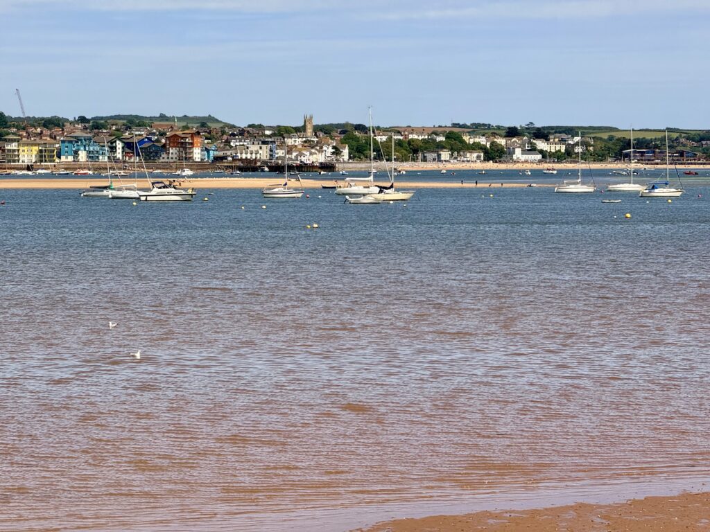 View across the Exe Estuary towards Exmouth, with sailing boats at anchor, the sandy shoreline and the East Devon seaside town in the background.