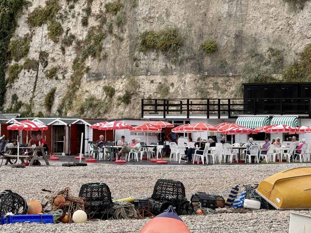 Ducky's Beach Café beneath the white chalk cliffs at Beer Beach, with outdoor tables, beach huts and fishing equipment on the shingle.