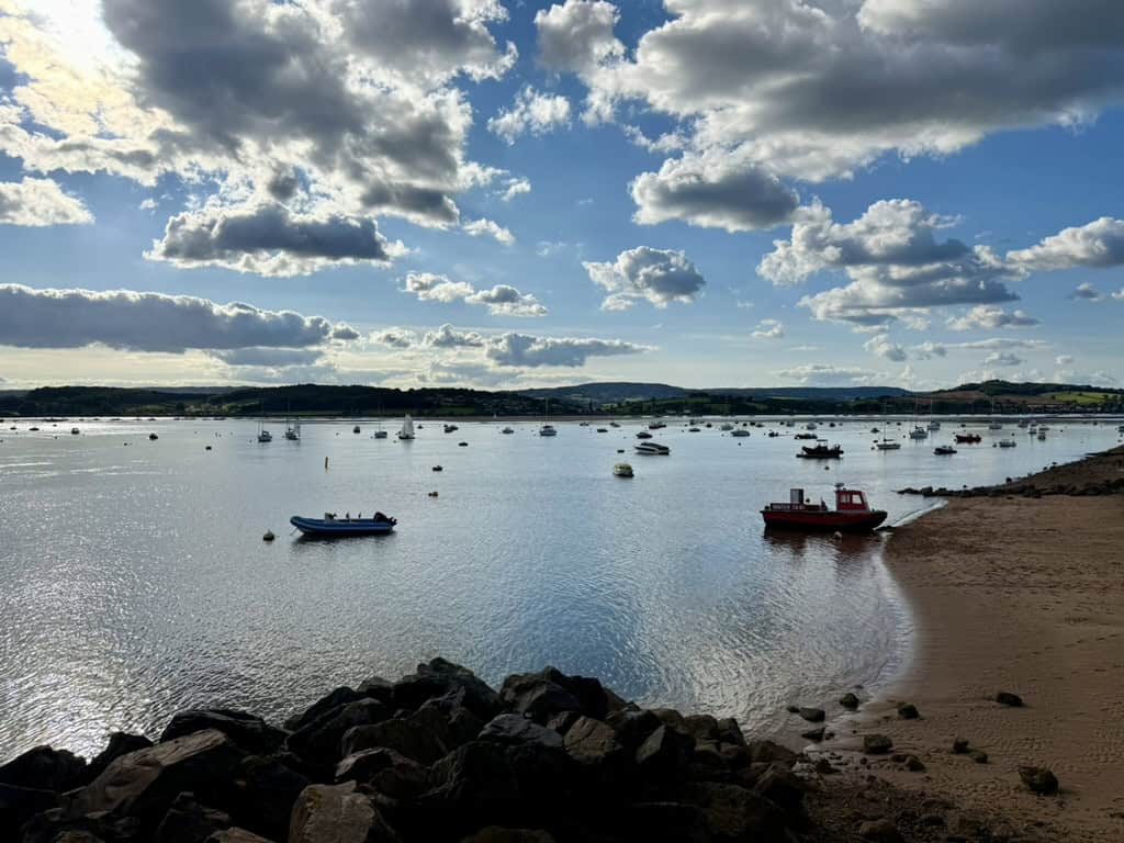 Shelley Beach (Pirate Cove), Exmouth, looking across the Exe Estuary towards Dawlish Warren with boats moored on calm water beneath dramatic clouds and late afternoon sunshine.