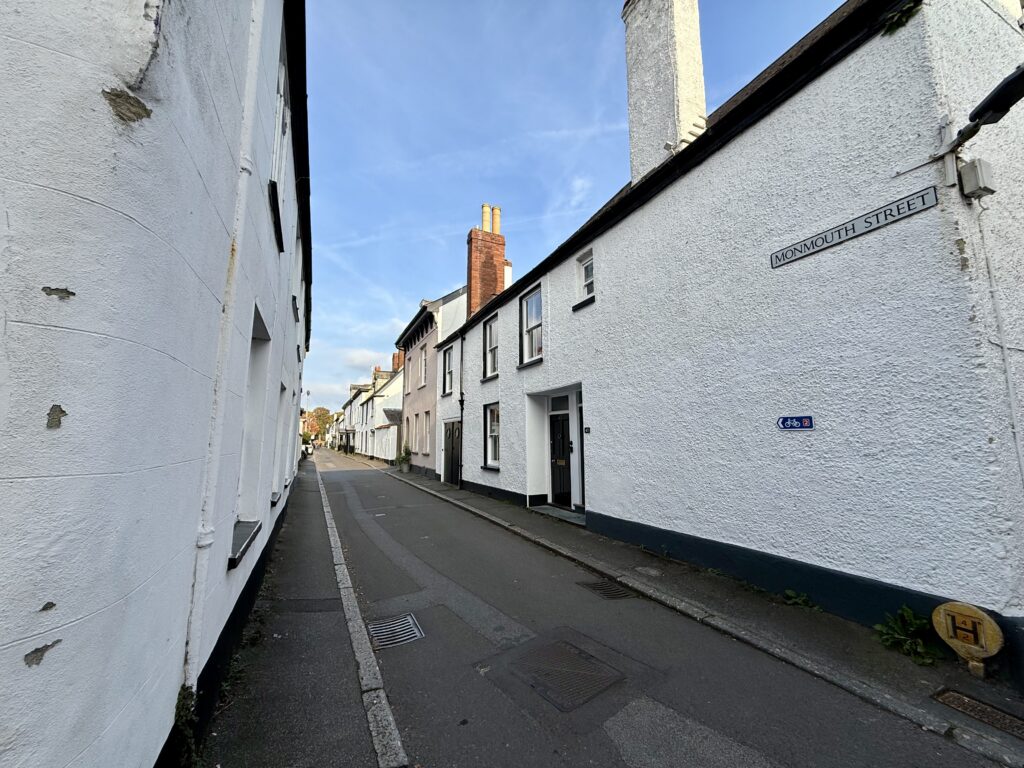 8FE85B7F-9B1E-4131-A468-E9CF5AB37020_1_201_a Monmouth Street in Topsham, with the Exe Estuary Trail cycle route passing between traditional whitewashed houses.