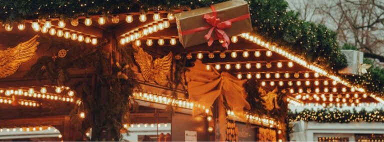 Festively decorated Christmas market stall adorned with warm string lights, evergreen garlands, golden angel ornaments, and a wrapped present with a red bow hanging from the roof.