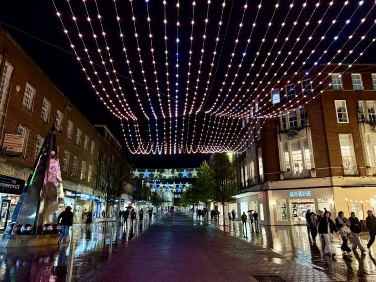Exeter High Street at night with festive Christmas lights hanging overhead, shoppers walking along the wet pavement and illuminated shopfronts creating a bright seasonal atmosphere.