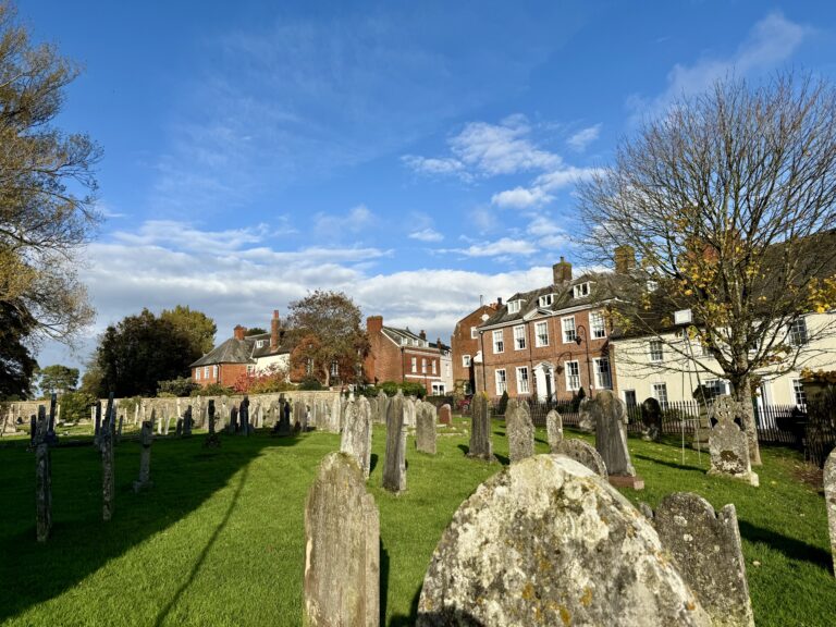 Churchyard at St Mary’s Church, Ottery St Mary, with old gravestones in the foreground and red-brick Georgian houses under a bright blue autumn sky.