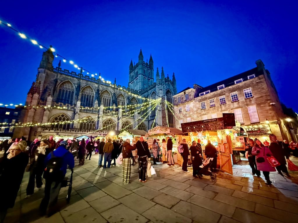 Bath Christmas Market at dusk, with illuminated wooden chalets, festive lights and visitors beside Bath Abbey