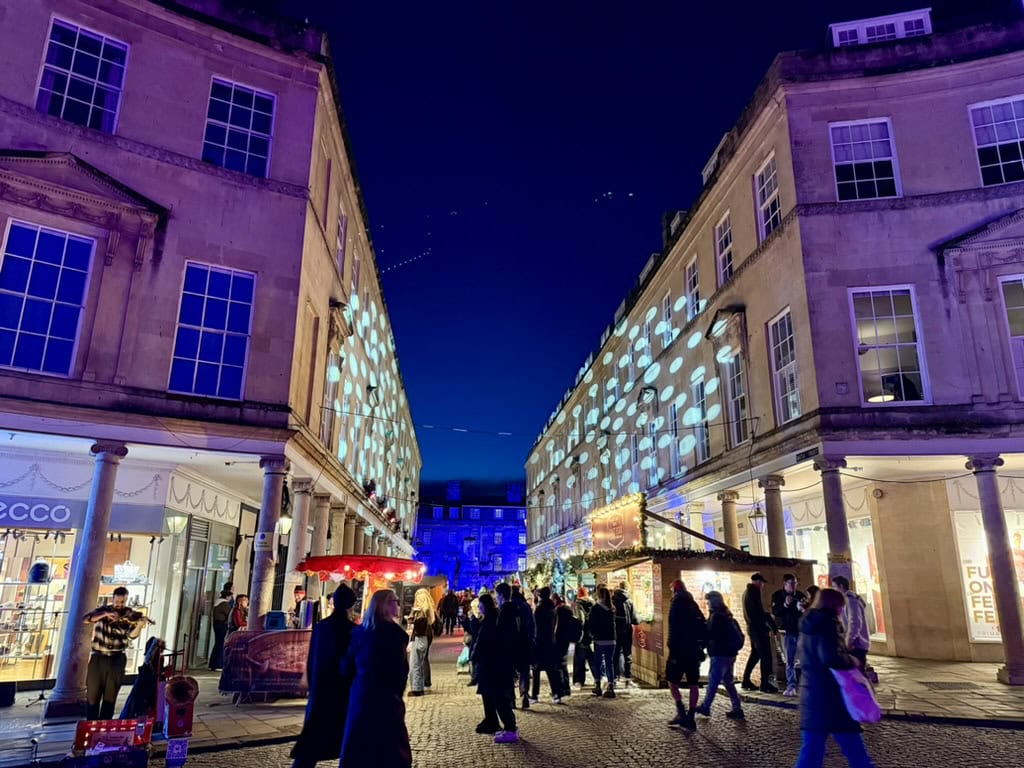 Bath Christmas Market at night with illuminated wooden chalets, festive light projections and visitors among Bath's Georgian streets