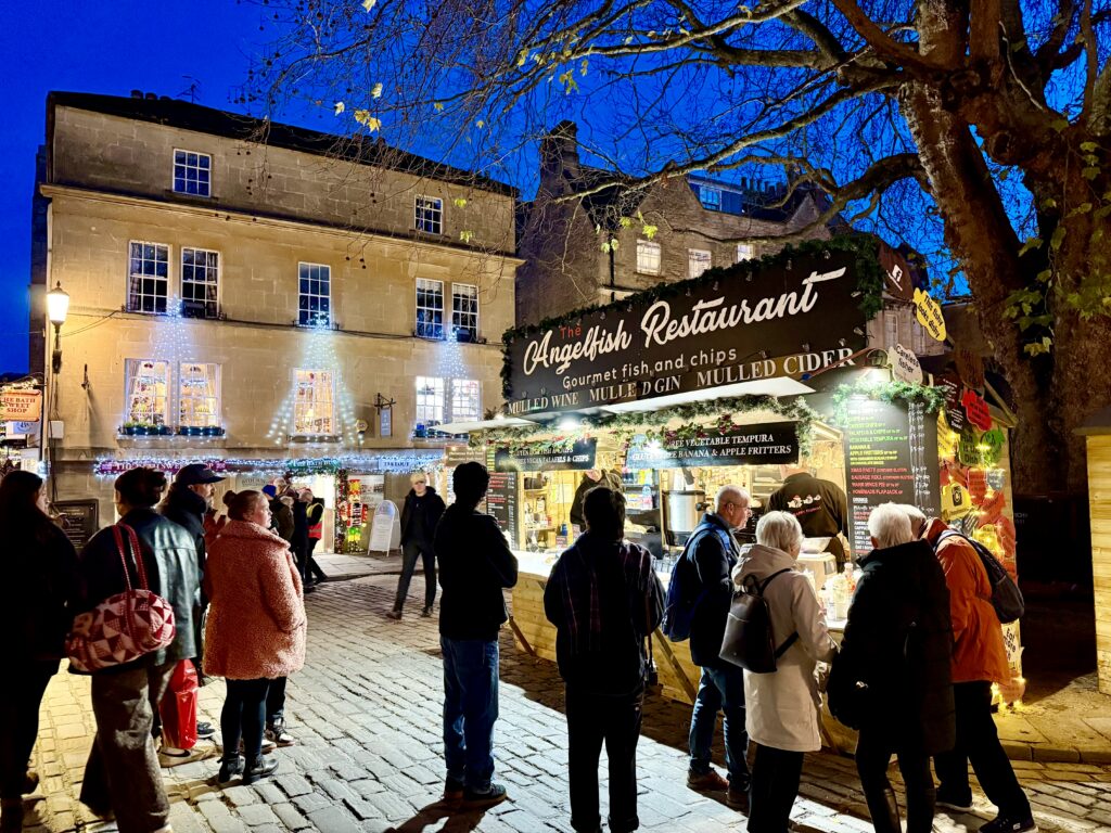 Visitors queue at an illuminated food stall serving fish and chips, mulled wine and cider at Bath Christmas Market