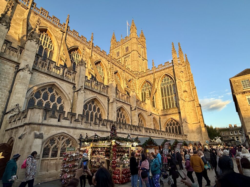Bath Christmas Market chalets and visitors beside the illuminated Gothic exterior of Bath Abbey at sunset.