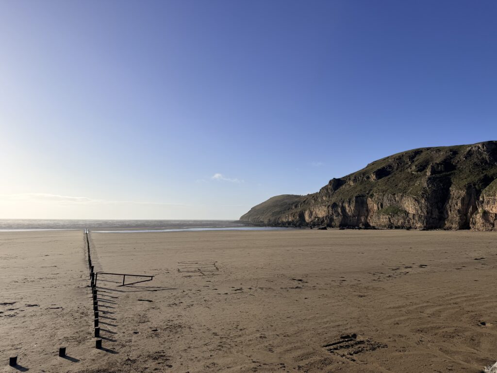 Brean Sands at low tide with wide sandy beach in the foreground and Brean Down rising to the right under a clear blue sky.