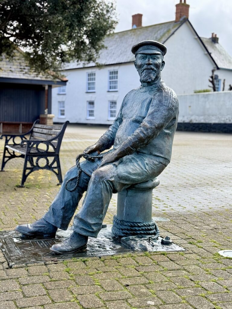 Yankee Jack statue in Watchet Somerset depicting a seated sailor with rope and cap, located near the harbour with historic buildings in the background.
