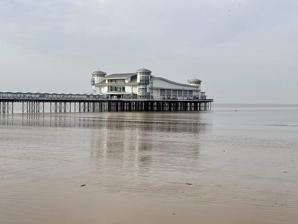 Ornate Victorian seafront shelter on the promenade in Weston-super-Mare with Weston Beach and the Grand Pier visible in the distance across Weston Bay on the Somerset coast.

