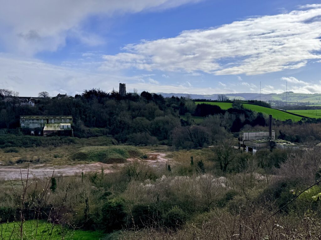 Wansbrough Paper Mill ruins near Watchet with St Decumans Church on the hillside behind, Somerset countryside landscape under a wide blue sky.
