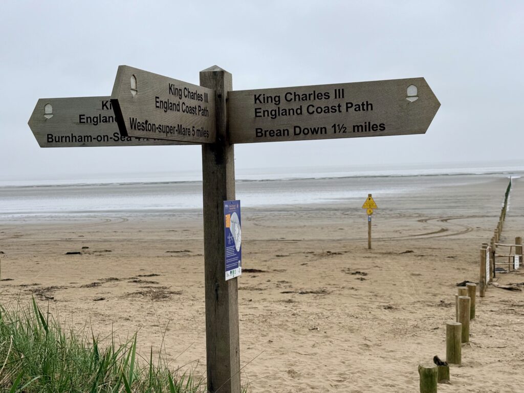 Signpost on Brean Beach showing directions for the King Charles III England Coast Path towards Brean Down, Weston-super-Mare and Burnham-on-Sea, with wide sandy beach and mudflats at low tide.
