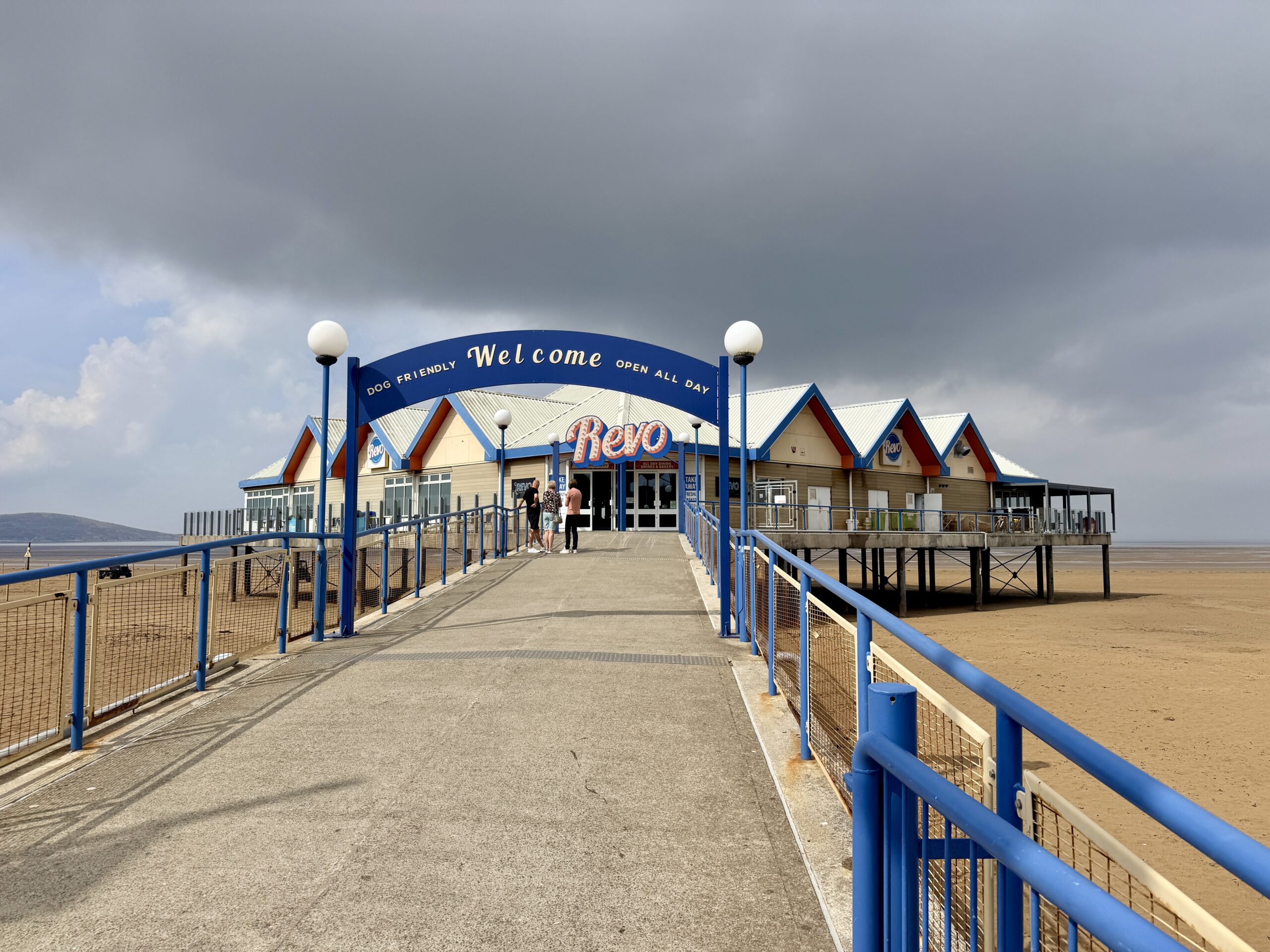 Revo Kitchen on Weston-super-Mare seafront, reached by a walkway over the beach, with Brean Down in the distance and dark storm clouds overhead.