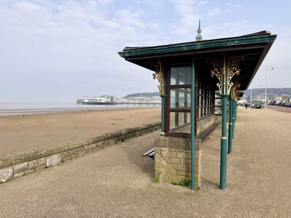 Decorative Victorian shelter on Weston-super-Mare promenade with Weston Beach and the Grand Pier visible across Weston Bay on the Somerset coast.