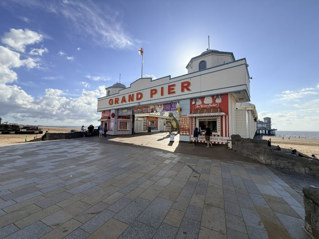 Grand Pier entrance on Weston-super-Mare seafront, with its distinctive red and white facade beside the wide paved promenade under a blue, partly cloudy sky.