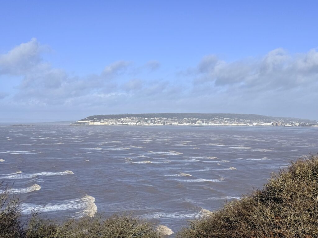 Panoramic view of Weston-super-Mare and Weston Bay across the Bristol Channel seen from Brean Down, showing the seaside town along the North Somerset coastline with waves rolling across the tidal waters.
