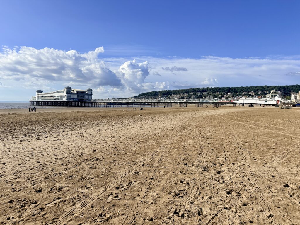 Wide sandy beach at Weston-super-Mare with the Grand Pier stretching across the bay and the wooded hillside and seafront buildings beyond.