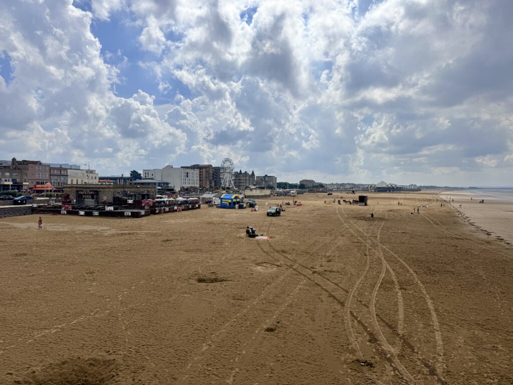 Weston-super-Mare's wide sandy beach at low tide, with seafront attractions, the big wheel beneath a dramatic cloudy sky.