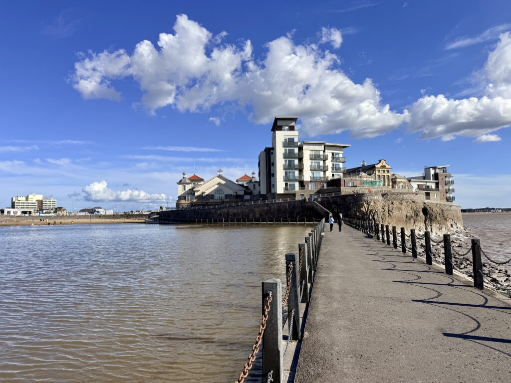 Causeway leading across Weston-super-Mare Marine Lake to Knightstone Island, with its historic buildings and modern apartments beneath a bright blue sky.
