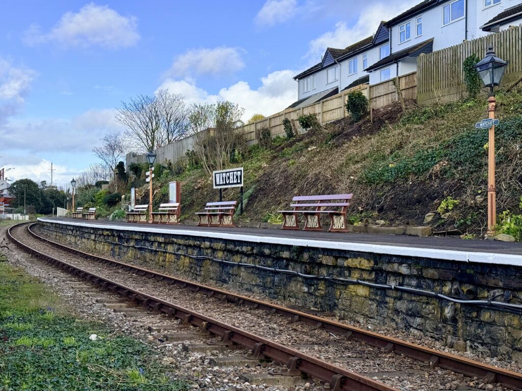 Watchet railway station platform on the West Somerset Railway with vintage signage, benches and track curving through the Somerset countryside.
