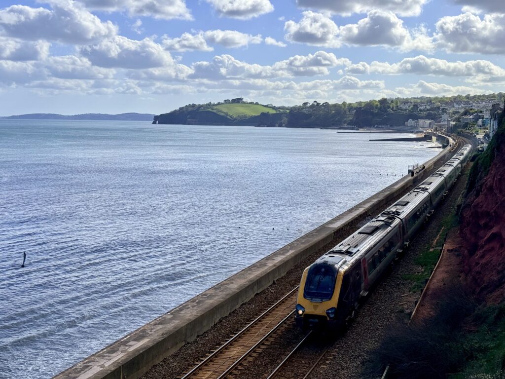 Train passing along the Dawlish sea wall in Devon with coastal views, red sandstone cliffs and the South Devon railway line beside the sea.