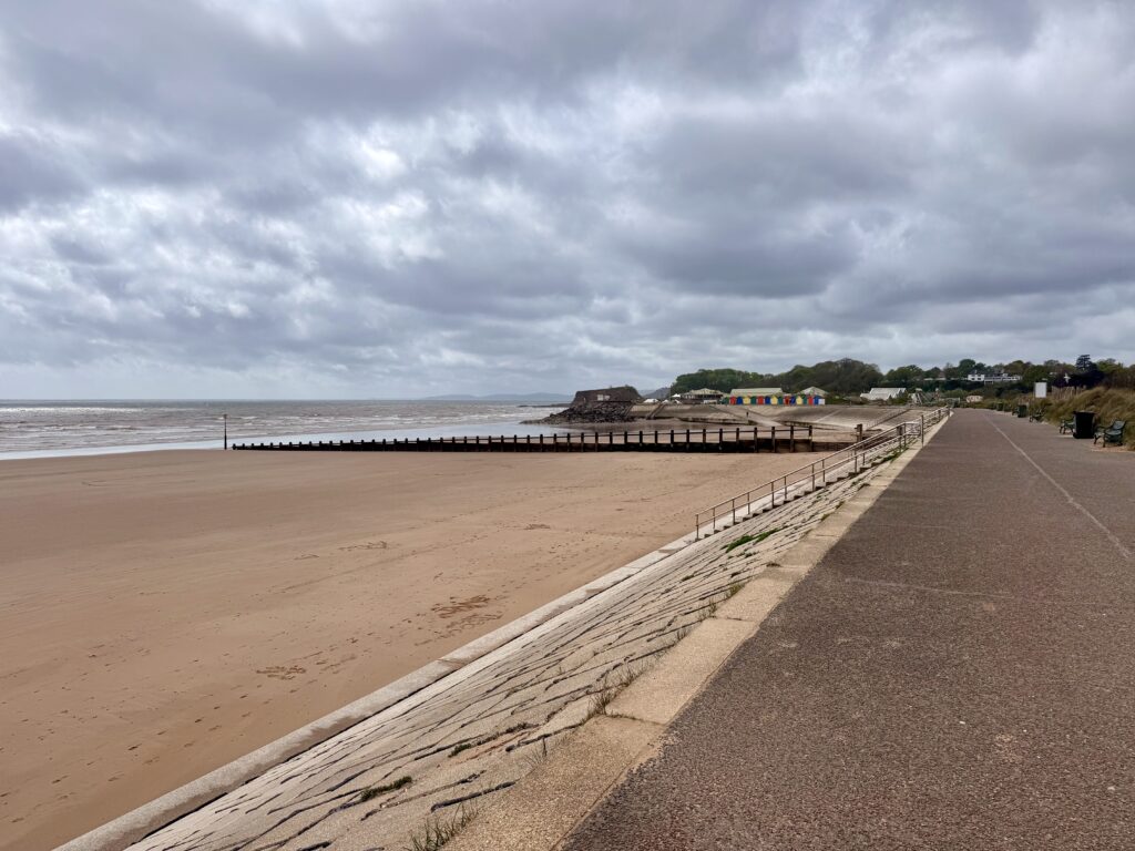 Dawlish Warren Beach, Devon with wide sandy shoreline, groynes, colourful beach huts and promenade under cloudy skies.