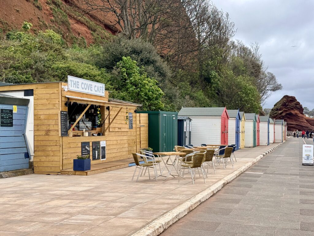 The Cove Café at Coryton Cove, Dawlish, Devon with colourful beach huts, outdoor seating and red sandstone cliffs behind.