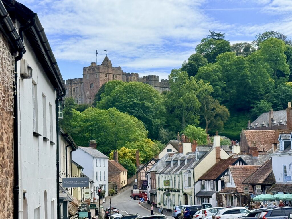 View of Dunster village with historic cottages and shops along the high street, with Dunster Castle rising above the trees on the hillside in Somerset.