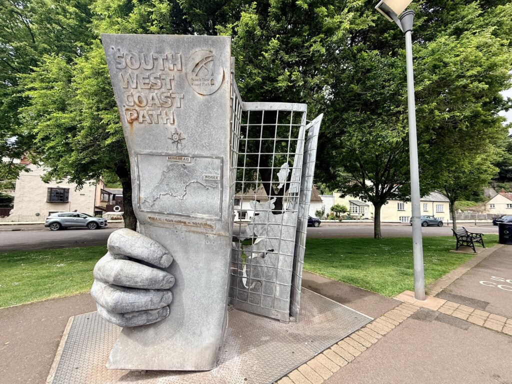 Sculpture marking the start of the South West Coast Path, one of the things to do in Minehead, showing large stone hands holding a map and guidebook, set beside the seafront path with trees and buildings nearby.