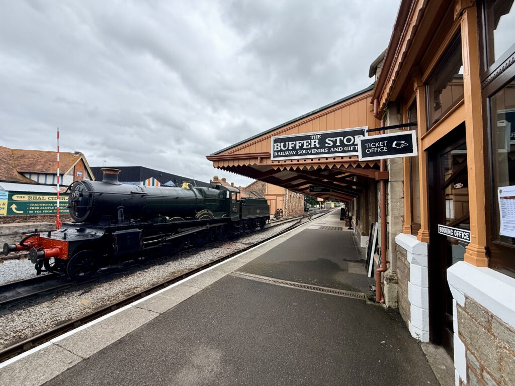 Things to do in Minehead include visiting Minehead railway station, where a black heritage steam locomotive stands at the platform beside a covered wooden station building, with signage for the West Somerset Railway and a quiet platform stretching into the distance.