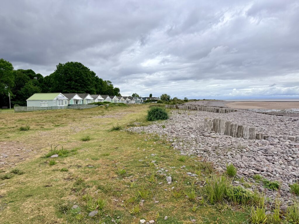 Watchet to Minehead walk at Dunster Beach with shingle shoreline, wooden groynes and coastal chalets, looking out across the Bristol Channel at low tide.