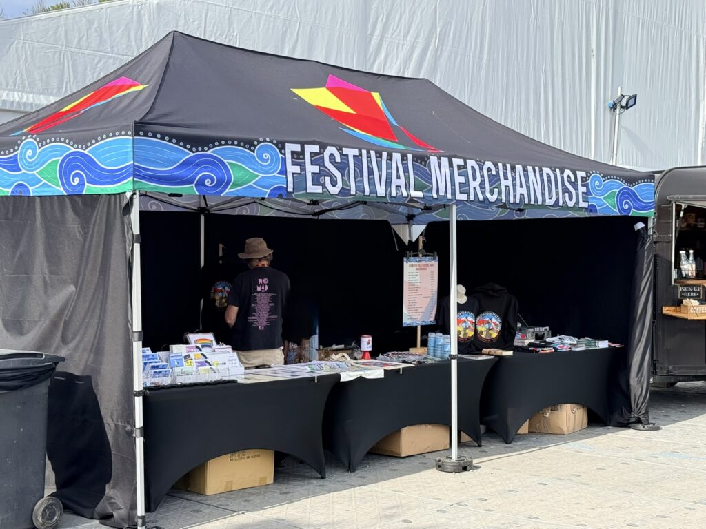 Sidmouth Folk Festival merchandise stall outside the Ham Marquee, displaying festival clothing, souvenirs and gifts in 2026