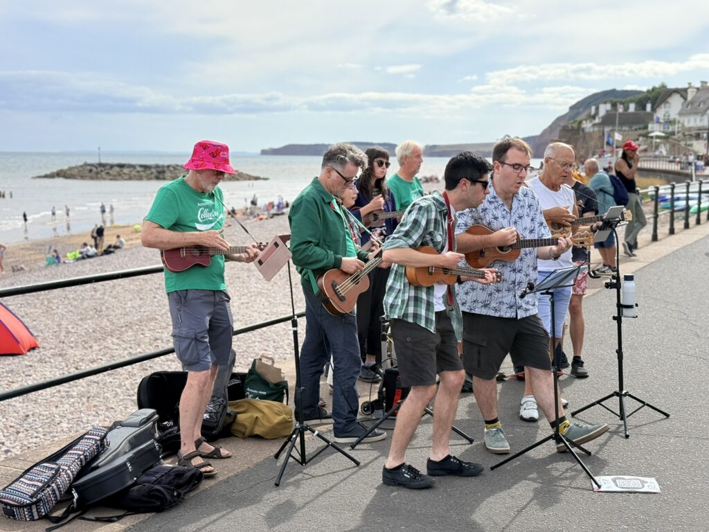 Group of musicians playing ukuleles on Sidmouth seafront during Sidmouth Folk Festival 2026, with the beach and sea behind them.