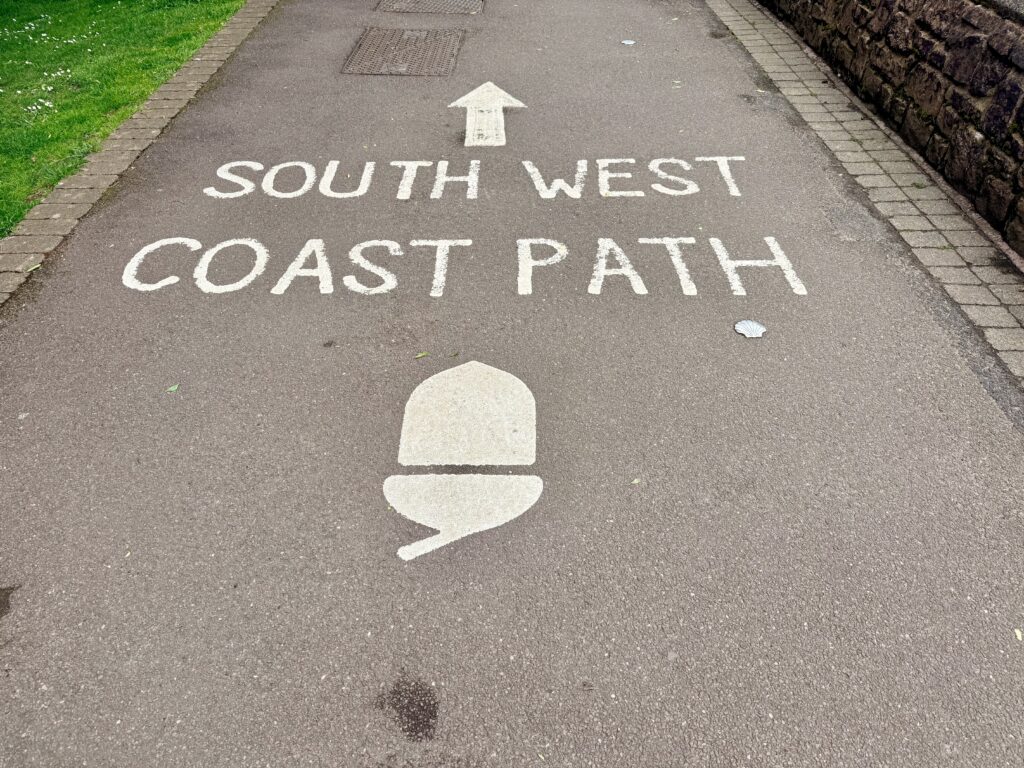 **Alt text:**
South West Coast Path sign painted on a promenade path in Minehead, Somerset, showing the acorn symbol marking the start of the South West Coast Path walking route.
