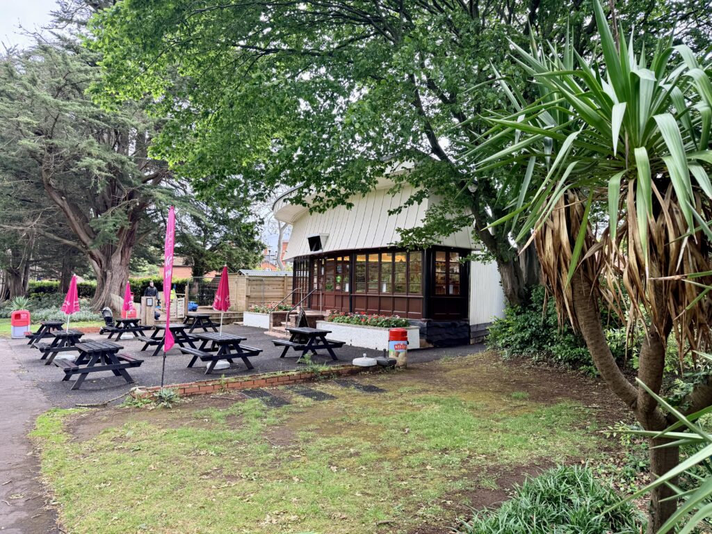 **Alt text:**
Blenheim Garden Café in Minehead, Somerset, with outdoor picnic tables, pink umbrellas and trees in the gardens near the seafront.