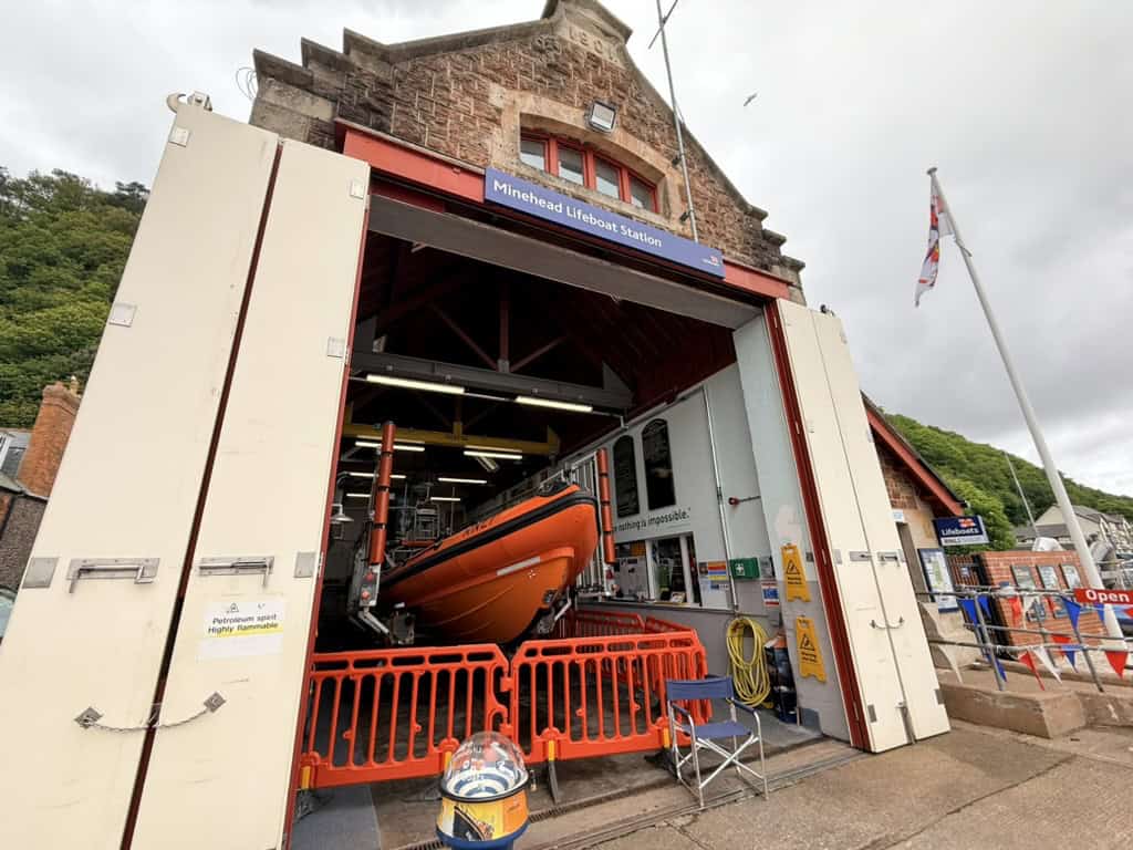 Visiting the RNLI lifeboat station in Minehead, showing an orange lifeboat inside an open boathouse on the harbour, with crew equipment and the station building behind.