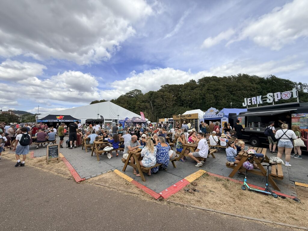 Visitors eating at outdoor picnic tables beside food stalls near the Ham Marquee at Sidmouth Folk Festival
