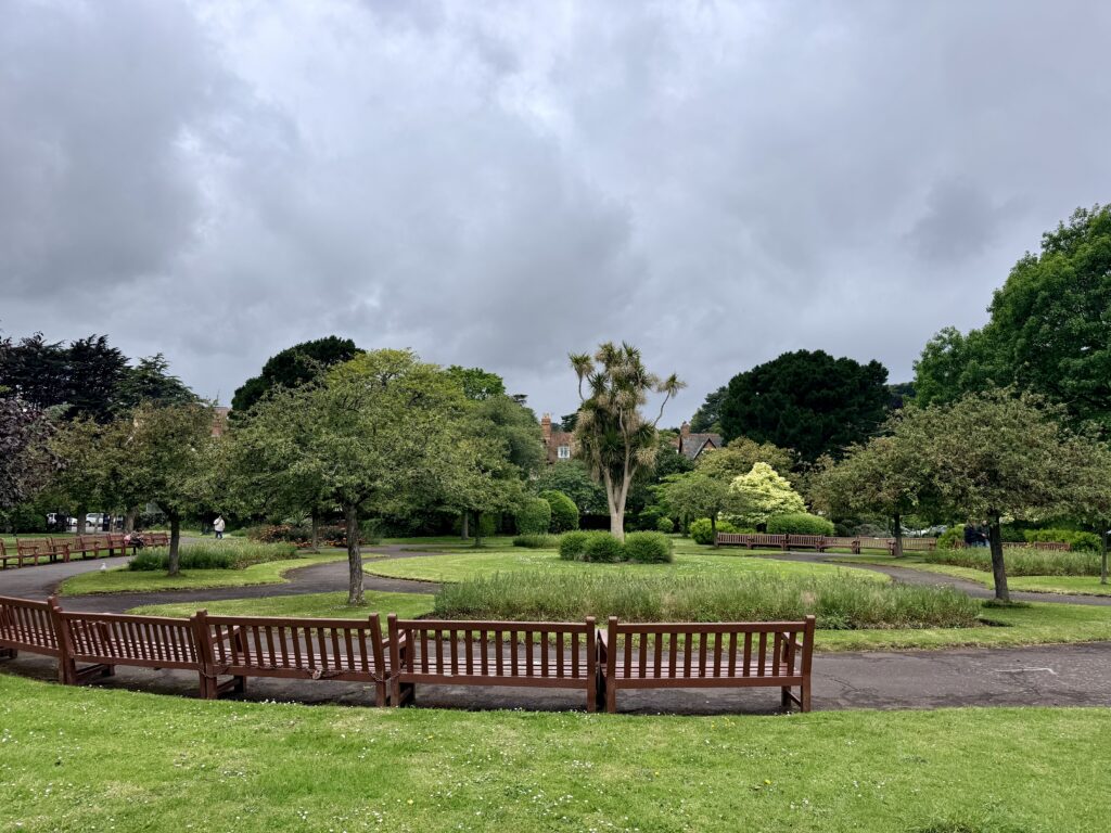 Blenheim Gardens in Minehead, Somerset, showing open lawns, mature trees, flower beds and wooden benches with footpaths through the park.