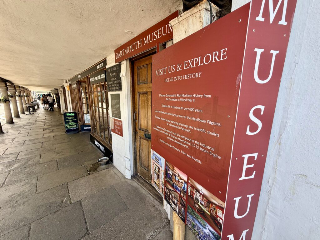 Entrance to Dartmouth Museum beneath The Butterwalk, with museum signs and displays along the covered pavement in Dartmouth town centre.