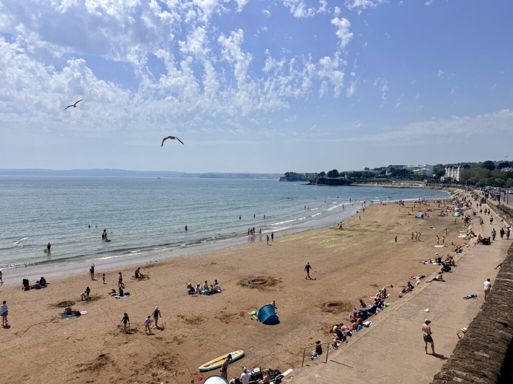 Torre Abbey Sands in Torquay showing the wide sandy beach, promenade and calm waters of Tor Bay on a busy summer day on the English Riviera.
