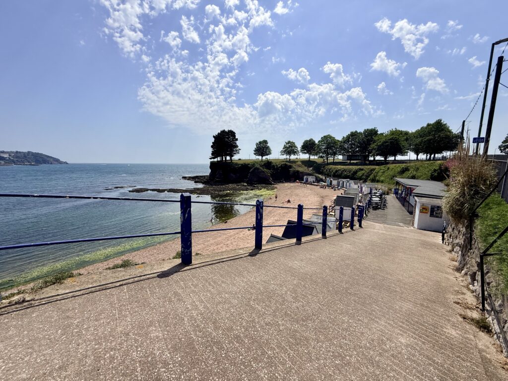 Corbyn Beach in Torquay with its sandy beach, colourful beach huts and Corbyn Head overlooking Tor Bay on a sunny day.