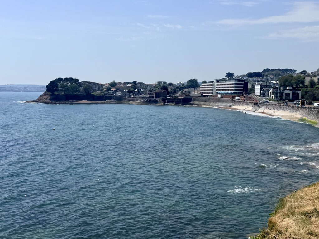 View across Livermead Sands in Torquay towards the Livermead Cliff Hotel with Institute Beach and the English Riviera coastline on a sunny day.