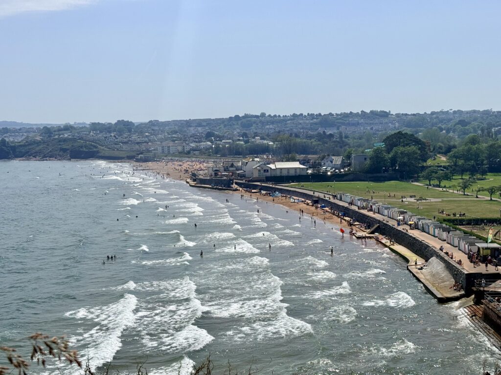 Goodrington Sands in Paignton showing the wide sandy beach, promenade, beach huts and Young's Park on the shores of Torbay on a sunny day.