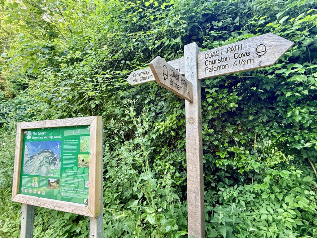 South West Coast Path signpost near Churston Cove showing directions to Brixham, Paignton and Greenway beside an information board in woodland on the English Riviera.