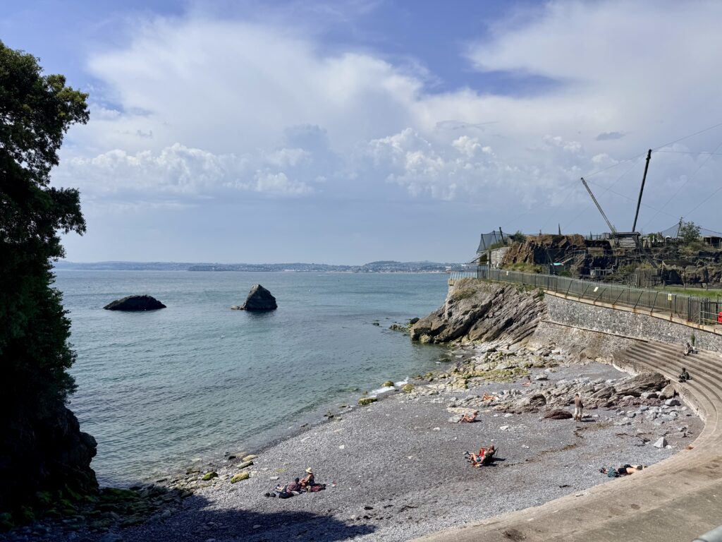 Beacon Cove in Torquay showing its shingle beach, rocky shoreline, clear water and sea defences beneath the Living Coasts headland on the English Riviera.