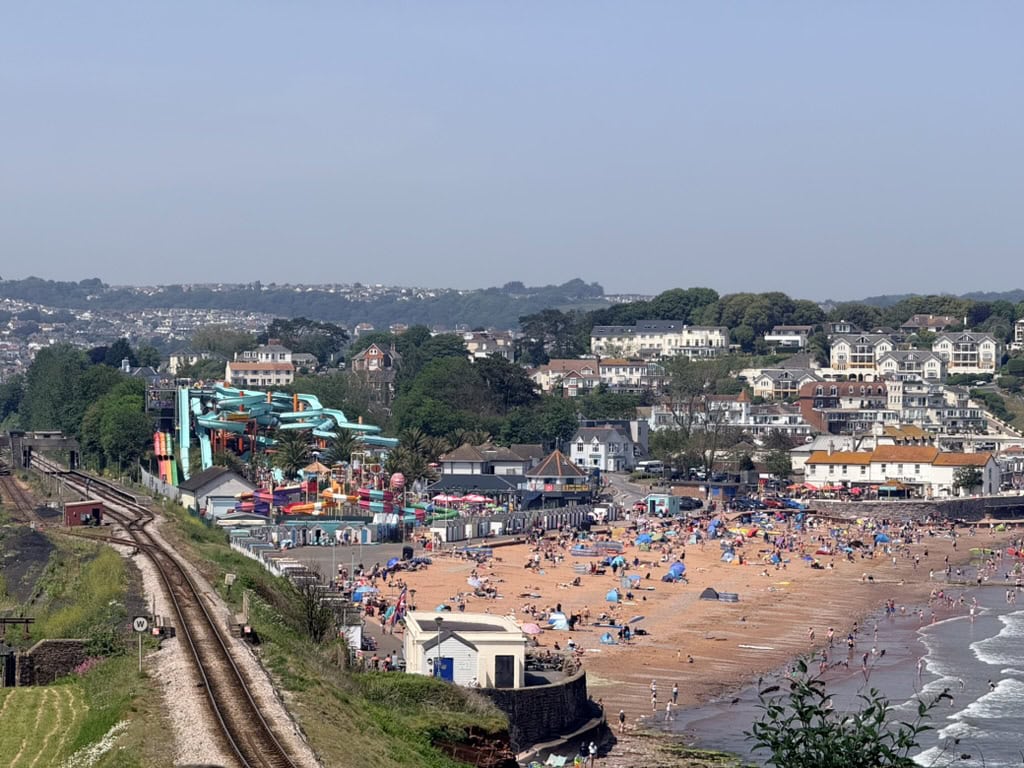 Goodrington Beach in Torbay, Devon, UK with families enjoying the sandy beach, Splashdown Quaywest waterpark, railway line and South Devon coastline in summer