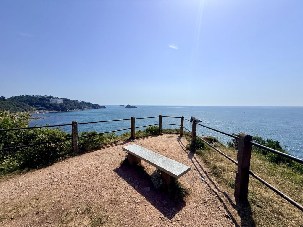Viewing point above Meadfoot Beach on the South West Coast Path overlooking Torbay with a bench, Thatcher Rock and the coastline towards Torquay on a sunny day.