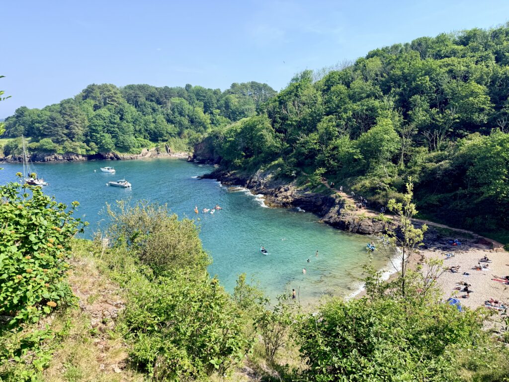Churston Cove near Brixham showing its secluded shingle beach, crystal clear water and wooded cliffs, with Fishcombe Cove visible in the distance on the English Riviera.