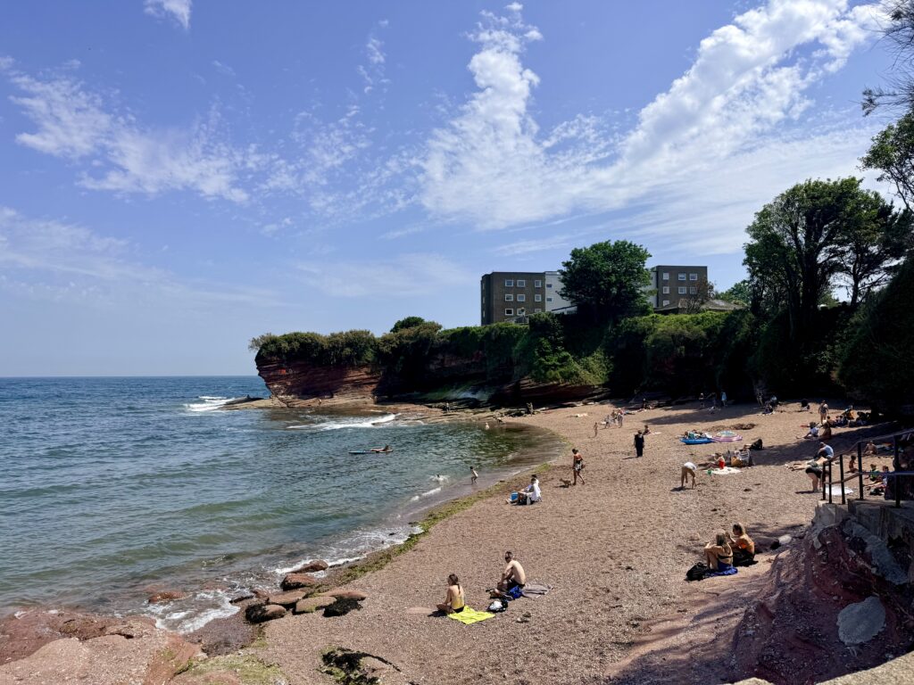 Fairy Cove in Paignton showing its sheltered sand and shingle beach beneath red sandstone cliffs on the shores of Torbay on a sunny day.