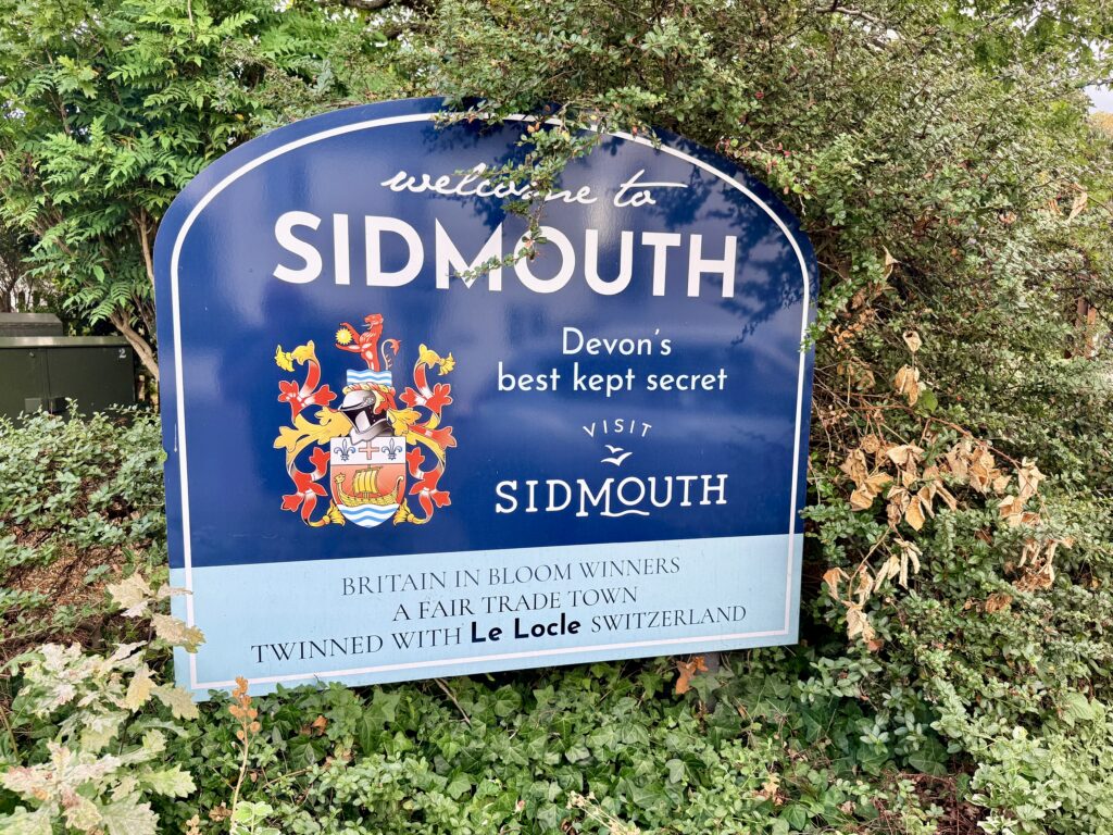 Welcome to Sidmouth sign surrounded by greenery, featuring the town crest and Visit Sidmouth branding in Devon.